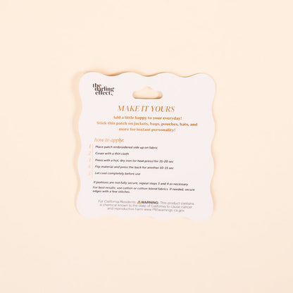 A square instruction card with scalloped edges and orange text, titled MAKE IT YOURS, guides you on using The Darling Effect’s Things Mix & Match Iron-On Patch to customize jackets. Includes steps, a warning, and website info at the bottom.