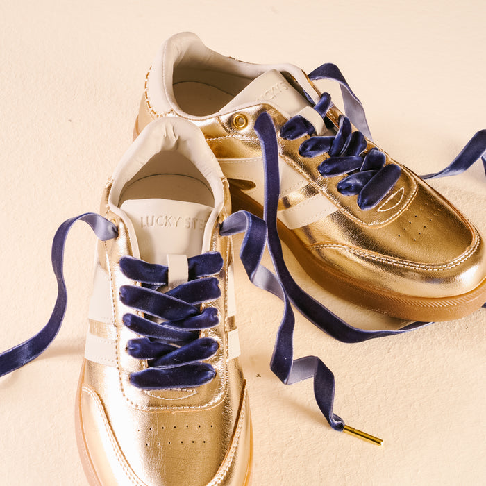 A pair of gold metallic sneakers with white accents and Velvet Blue Step It Up Shoelaces by The Darling Effect, displayed on a light beige surface. The inside of one shoe shows the brand name LUCKY STEP.
