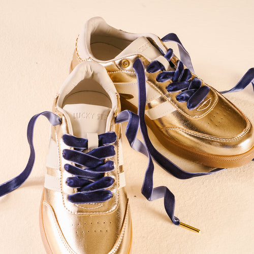 A pair of gold metallic sneakers with white accents and Velvet Blue Step It Up Shoelaces by The Darling Effect, displayed on a light beige surface. The inside of one shoe shows the brand name LUCKY STEP.