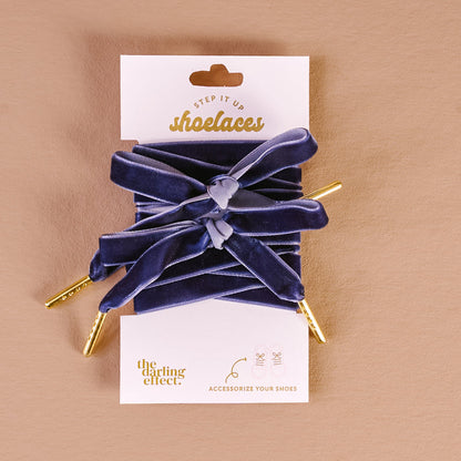 A pair of wide, stylish velvet blue shoelaces with gold tips are tied in bows and displayed on a branded card reading Velvet Blue Step It Up Shoelaces by The Darling Effect against a beige background.
