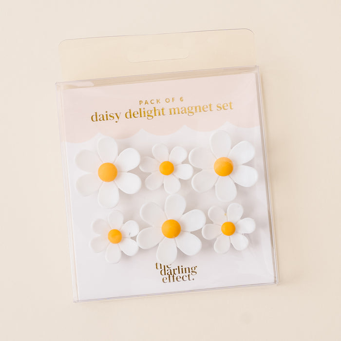 The White Daisy Delight Magnet Set by the-darling-effect features six vibrant daisy-shaped magnets with white petals and yellow centers in clear packaging, providing a strong hold against a light cream background.