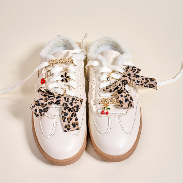 The Darling Effect's Wild & Free Dazzle Shoe Charm Pack decorates white sneakers featuring light brown soles, fuzzy lining, leopard print bows, gold chain accents, and cherry charms, all showcased on a neutral background.