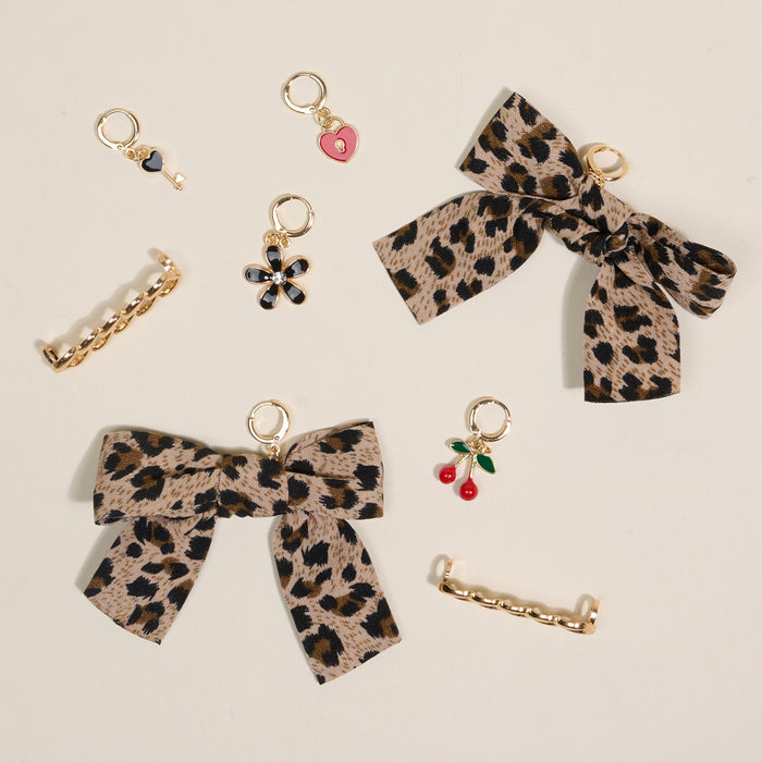 The Darling Effect's Wild & Free Dazzle Shoe Charm Pack includes two leopard print bows, two gold hair clips, five small gold charms (heart, flower, cherries, pearl), and sneaker charms—arranged on a light background.