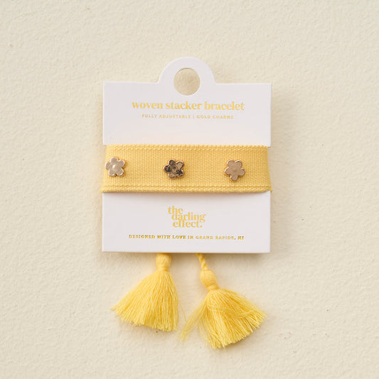 The Darling Effect’s Yellow Woven Stacker Bracelet features gold flower details and tassels, displayed on a branded card. Designed with love in Grand Rapids, MI—perfect for fans of adjustable bracelets.