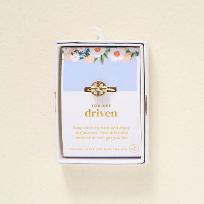 The Darling Effect's You Are Driven Adjustable Ring features a geometric 14k gold design, presented in a white box. Behind it, a floral card reads "You are driven" and shares an inspiring message about hard work and dedication.