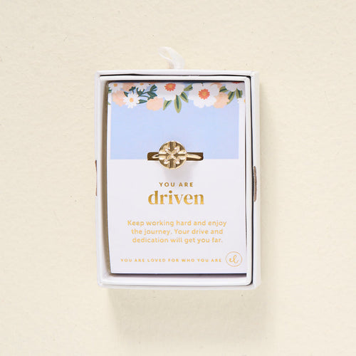 The Darling Effect's You Are Driven Adjustable Ring features a geometric 14k gold design, presented in a white box. Behind it, a floral card reads "You are driven" and shares an inspiring message about hard work and dedication.
