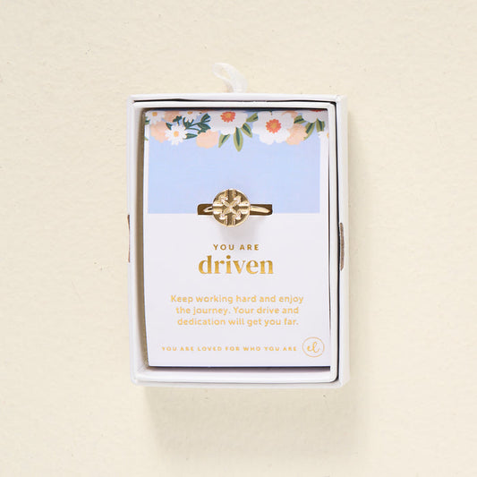 The Darling Effect's You Are Driven Adjustable Ring features a geometric 14k gold design, presented in a white box. Behind it, a floral card reads "You are driven" and shares an inspiring message about hard work and dedication.